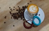 A glass of latte with intricate latte art on top sits on a white saucer, surrounded by scattered coffee beans on a light wooden surface. Two coiled wrist lanyards, one red and one blue, rest beside the glass.
