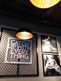 Industrial-style interior featuring a metal mesh wall art with text 'Catch up with everything you love'. Two hanging pendant lights with warm illumination. Adjacent black and white framed photographs depict a person holding a coffee cup.
