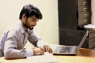A man with dark hair and a beard is writing in a notebook while seated at a wooden desk. He is focused on his task, using a pen, and has a laptop open next to him. In the background, there is a set of drawers and some papers stacked.