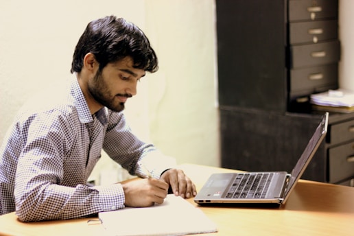 A man with dark hair and a beard is writing in a notebook while seated at a wooden desk. He is focused on his task, using a pen, and has a laptop open next to him. In the background, there is a set of drawers and some papers stacked.