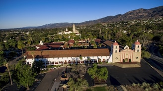 A panoramic view of a large mission-style building with distinct red rooftops and arched windows, surrounded by lush greenery and trees. The structure is situated in a picturesque location with mountains in the background, under a clear blue sky.