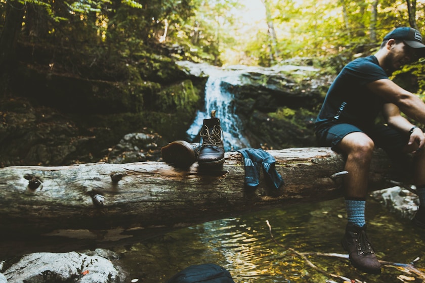 man sitting on tree log beside boots break