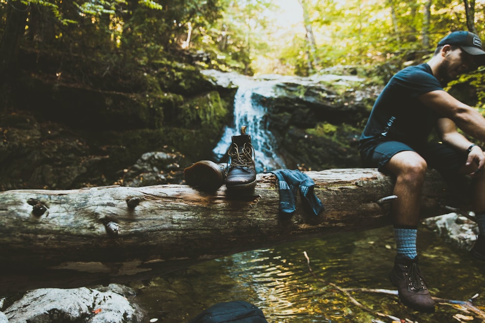 man sitting on tree log beside boots