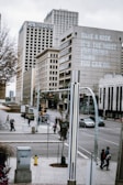 A city street scene with tall office buildings, including one with the words 'Take a risk. It's the most educational thing you can do.' Pedestrians walk along the sidewalks and cross the intersection, while vehicles are on the road. There are trees and urban street fixtures, under a cloudy sky.