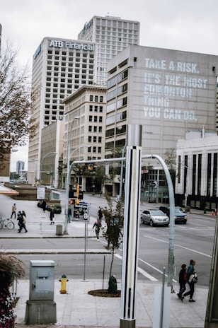 A city street scene with tall office buildings, including one with the words 'Take a risk. It's the most educational thing you can do.' Pedestrians walk along the sidewalks and cross the intersection, while vehicles are on the road. There are trees and urban street fixtures, under a cloudy sky.