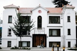 A white, multi-story building with brown trim features a sign that reads 'El Mirador 10147'. The structure has a tiled roof and several windows, with a tree partially obscuring its facade. A person walks on the sidewalk in front of the building, and a lamp post is visible to the right.