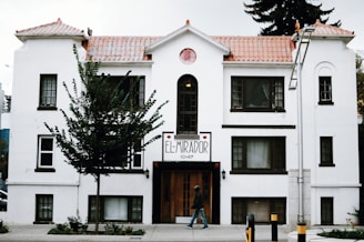 A white, multi-story building with brown trim features a sign that reads 'El Mirador 10147'. The structure has a tiled roof and several windows, with a tree partially obscuring its facade. A person walks on the sidewalk in front of the building, and a lamp post is visible to the right.
