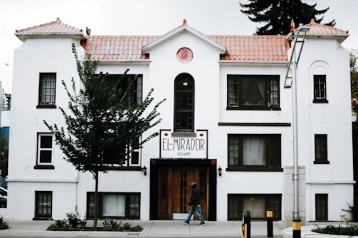 A white, multi-story building with brown trim features a sign that reads 'El Mirador 10147'. The structure has a tiled roof and several windows, with a tree partially obscuring its facade. A person walks on the sidewalk in front of the building, and a lamp post is visible to the right.