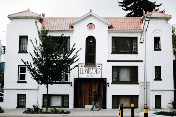 A white, multi-story building with brown trim features a sign that reads 'El Mirador 10147'. The structure has a tiled roof and several windows, with a tree partially obscuring its facade. A person walks on the sidewalk in front of the building, and a lamp post is visible to the right.