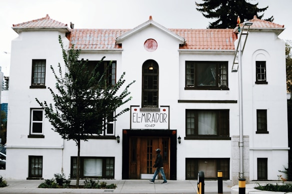 A white, multi-story building with brown trim features a sign that reads 'El Mirador 10147'. The structure has a tiled roof and several windows, with a tree partially obscuring its facade. A person walks on the sidewalk in front of the building, and a lamp post is visible to the right.
