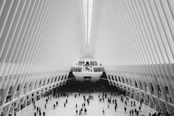 A modern architectural space features a high, vaulted ceiling with symmetrical, rib-like beams creating a sense of expansive openness. A crowd of people are scattered across the spacious floor area, giving a dynamic sense of movement and bustle within the grandeur of the structure.