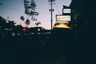 A cozy taquería storefront near a busy city monument at dusk.