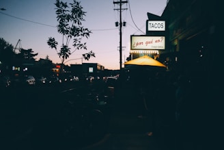 A city street scene during twilight, with a line of cars visible and the street illuminated by car headlights. A neon sign displaying the word 'TACOS' hangs prominently on a building, accompanied by another sign saying '¿por qué no? Taqueria'. An outdoor seating area with a yellow umbrella is also visible.