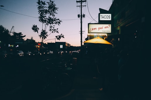A city street scene during twilight, with a line of cars visible and the street illuminated by car headlights. A neon sign displaying the word 'TACOS' hangs prominently on a building, accompanied by another sign saying '¿por qué no? Taqueria'. An outdoor seating area with a yellow umbrella is also visible.