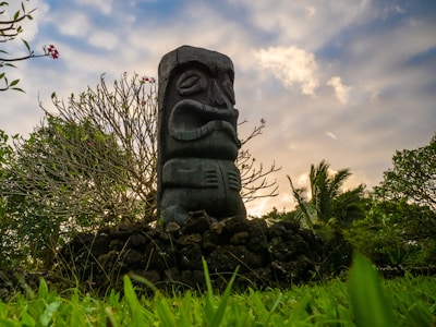 A large stone tiki statue stands on a mound of lava rocks, surrounded by lush green grass and tropical foliage. The sky behind is partly cloudy, casting a warm evening glow over the scene, and a few small red flowers are visible on a tree to the left.