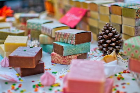 A variety of handmade soap bars are artfully arranged on a table. Each soap is wrapped in colorful paper with different patterns and colors. Scattered small decorative candies and a pine cone add to the festive atmosphere. The soaps vary in color, including brown, pink, and yellow hues, and are placed alongside small meringue-like decorations.