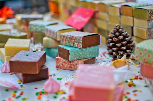 A variety of handmade soap bars are artfully arranged on a table. Each soap is wrapped in colorful paper with different patterns and colors. Scattered small decorative candies and a pine cone add to the festive atmosphere. The soaps vary in color, including brown, pink, and yellow hues, and are placed alongside small meringue-like decorations.