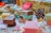 A variety of handmade soap bars are artfully arranged on a table. Each soap is wrapped in colorful paper with different patterns and colors. Scattered small decorative candies and a pine cone add to the festive atmosphere. The soaps vary in color, including brown, pink, and yellow hues, and are placed alongside small meringue-like decorations.