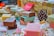 A variety of handmade soap bars are artfully arranged on a table. Each soap is wrapped in colorful paper with different patterns and colors. Scattered small decorative candies and a pine cone add to the festive atmosphere. The soaps vary in color, including brown, pink, and yellow hues, and are placed alongside small meringue-like decorations.