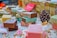 A variety of handmade soap bars are artfully arranged on a table. Each soap is wrapped in colorful paper with different patterns and colors. Scattered small decorative candies and a pine cone add to the festive atmosphere. The soaps vary in color, including brown, pink, and yellow hues, and are placed alongside small meringue-like decorations.