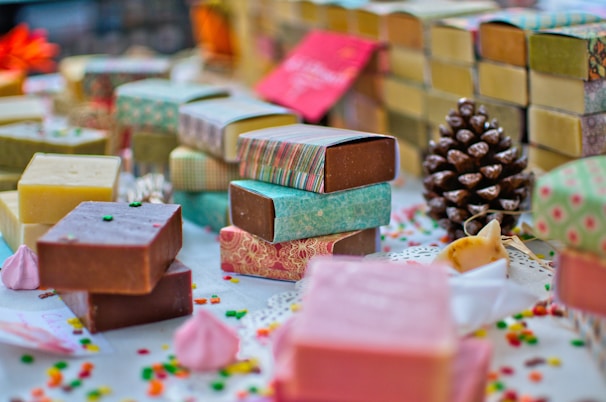 A variety of handmade soap bars are artfully arranged on a table. Each soap is wrapped in colorful paper with different patterns and colors. Scattered small decorative candies and a pine cone add to the festive atmosphere. The soaps vary in color, including brown, pink, and yellow hues, and are placed alongside small meringue-like decorations.