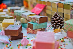 A variety of handmade soap bars are artfully arranged on a table. Each soap is wrapped in colorful paper with different patterns and colors. Scattered small decorative candies and a pine cone add to the festive atmosphere. The soaps vary in color, including brown, pink, and yellow hues, and are placed alongside small meringue-like decorations.
