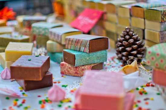 A variety of handmade soap bars are artfully arranged on a table. Each soap is wrapped in colorful paper with different patterns and colors. Scattered small decorative candies and a pine cone add to the festive atmosphere. The soaps vary in color, including brown, pink, and yellow hues, and are placed alongside small meringue-like decorations.