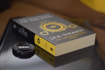 A paperback book titled 'The Lord of the Rings' by J.R.R. Tolkien rests on a reflective surface alongside a Nikon camera lens cap. The book cover is primarily dark with gold circular designs, and the side displays the author's name and book title in gold letters.