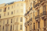 A view of European-style residential buildings featuring several stories and ornate wrought iron balconies. The architecture is classical with a warm, sunlit yellow hue casting a soft glow. The windows are tall and rectangular, designed with decorative elements.