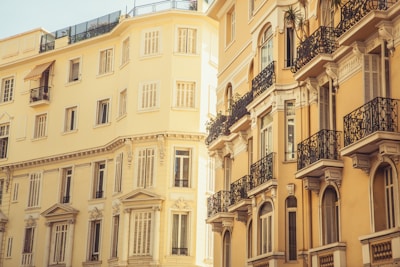 A view of European-style residential buildings featuring several stories and ornate wrought iron balconies. The architecture is classical with a warm, sunlit yellow hue casting a soft glow. The windows are tall and rectangular, designed with decorative elements.