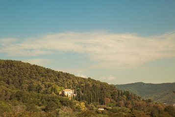 A serene landscape showcasing a house nestled amidst lush greenery, surrounded by rolling hills covered in dense forests under a clear blue sky.