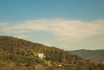 A serene landscape showcasing a house nestled amidst lush greenery, surrounded by rolling hills covered in dense forests under a clear blue sky.