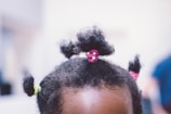 The image features the top of a child's head with several small, curly hair buns secured with colorful hair ties in pink and green.