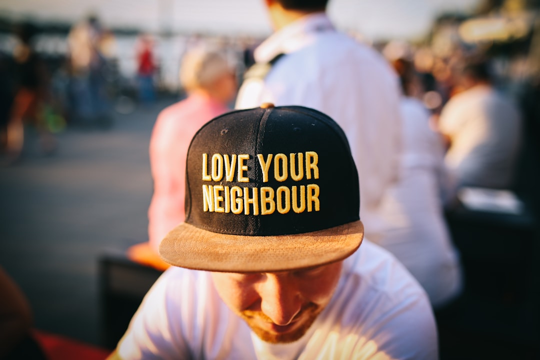 A man wears a hat that reads "Love Your Neighbour" on crowded Rheinpromenade