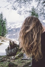 Long, wavy blonde hair flows down against a backdrop of a mountainous landscape with snow patches and tall evergreen trees. A fallen tree is visible, with its roots exposed, surrounded by patches of grass and snow.