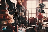 A small, dimly lit vintage shop filled with an eclectic assortment of items including a variety of hats, bags, and decorative objects. The shop has wooden shelves and cabinets, with hats displayed prominently. A chair sits near a sunlit window, adding a warm glow to the scene.