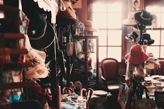 A small, dimly lit vintage shop filled with an eclectic assortment of items including a variety of hats, bags, and decorative objects. The shop has wooden shelves and cabinets, with hats displayed prominently. A chair sits near a sunlit window, adding a warm glow to the scene.