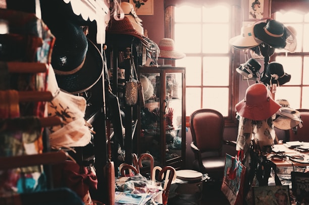 A small, dimly lit vintage shop filled with an eclectic assortment of items including a variety of hats, bags, and decorative objects. The shop has wooden shelves and cabinets, with hats displayed prominently. A chair sits near a sunlit window, adding a warm glow to the scene.