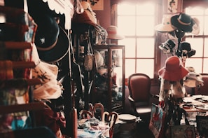 A small, dimly lit vintage shop filled with an eclectic assortment of items including a variety of hats, bags, and decorative objects. The shop has wooden shelves and cabinets, with hats displayed prominently. A chair sits near a sunlit window, adding a warm glow to the scene.