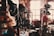 A small, dimly lit vintage shop filled with an eclectic assortment of items including a variety of hats, bags, and decorative objects. The shop has wooden shelves and cabinets, with hats displayed prominently. A chair sits near a sunlit window, adding a warm glow to the scene.