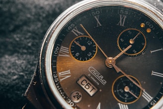 A close-up view of an analog wristwatch with a dark face, Roman numerals, and multiple small dials. The watch includes gold-colored hands and markings, with visible indicators for the day and date. The background is black and textured, enhancing the vintage feel.