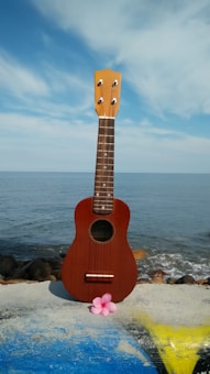 A wooden ukulele is placed upright on a concrete surface, with the sea and a blue sky in the background. Pink flower petals are laid in front of the ukulele, adding a touch of color to the scene.