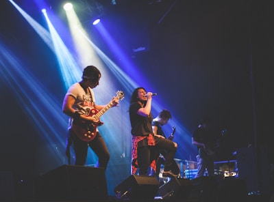 A band performs on stage with vibrant lighting effects. The lead singer, holding a microphone and wearing casual attire with a plaid shirt tied around the waist, sings passionately into the microphone. Other band members play instruments, including electric guitars. The background features colorful stage lights creating a dynamic atmosphere.