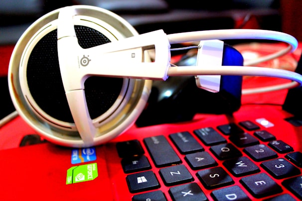 A pair of large white headphones rests on the keyboard of a red laptop. The laptop's keys are black with white lettering, and there are visible stickers indicating an Intel processor and NVIDIA graphics. The focus is on the headphones, which have a thick headband and large ear cups.
