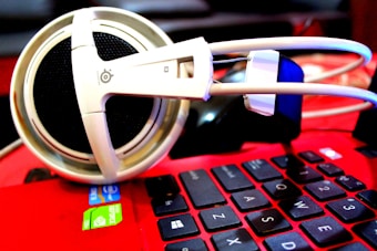 A pair of large white headphones rests on the keyboard of a red laptop. The laptop's keys are black with white lettering, and there are visible stickers indicating an Intel processor and NVIDIA graphics. The focus is on the headphones, which have a thick headband and large ear cups.