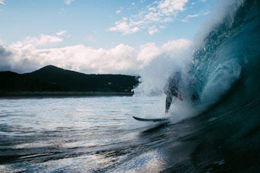 A surfer rides a large wave, partially obscured by the crashing water. The scene captures the dynamic motion of the wave and the surfer's skillful maneuvering. A coastline with mountains is visible in the background under a partly cloudy sky.