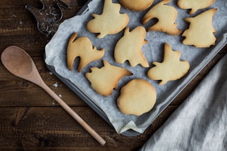 A tray of freshly baked cookies in various Halloween-themed shapes, resting on a piece of parchment paper. A wooden spoon lies nearby on a rustic wooden surface, while a metal cookie cutter is visible near some scattered flour. A textured, grey cloth complements the cozy and inviting baking scene.