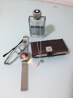 A wristwatch with a metallic mesh strap lies next to a pair of reading glasses. Nearby, there's a closed dark leather wallet and a sleek, square-shaped bottle. A silver chain and pendant are draped across the bottle and wallet.