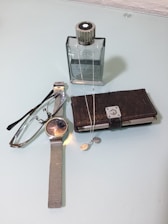 A wristwatch with a metallic mesh strap lies next to a pair of reading glasses. Nearby, there's a closed dark leather wallet and a sleek, square-shaped bottle. A silver chain and pendant are draped across the bottle and wallet.