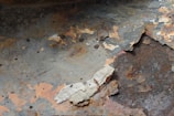 Peeling and rusted metal surface with layers of paint flaking off. The underlying rust and corrosion are visible, showing various shades of brown and orange. There are patches where the paint has completely peeled away, exposing the raw metal. The texture is rough and uneven.